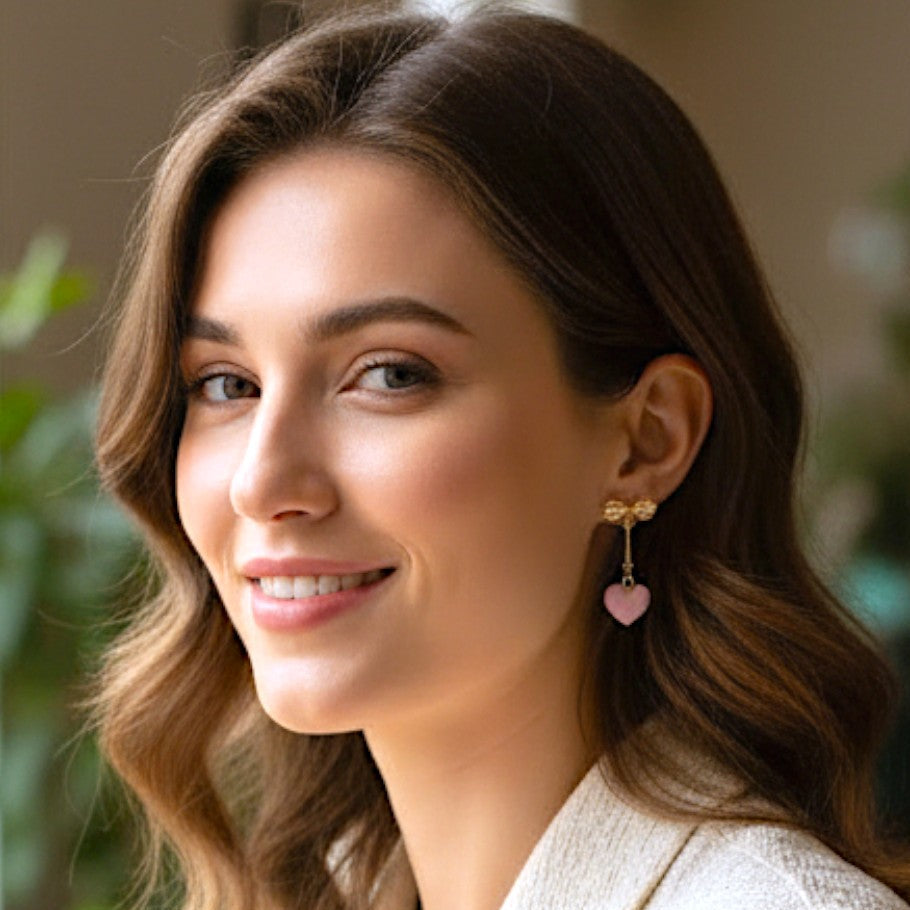 Woman wearing a pair of earrings with a pink, heart hanging from a gold bow with a indoor background.