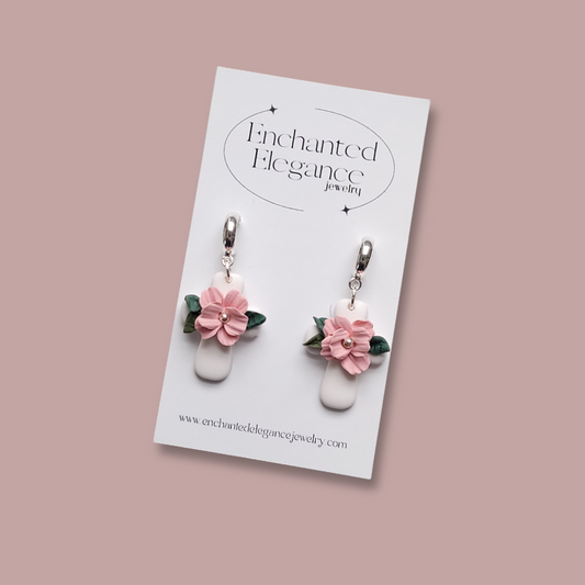 Hand-sculpted white polymer clay cross earrings with a pink flower and green leaves on silver hoops, displayed on an Enchanted Elegance branded card