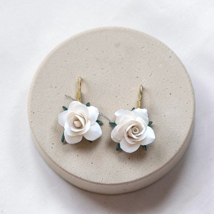 Hand-sculpted white polymer clay rose earrings with tiny green leaves, displayed on a round concrete jewelry pedestal against white linen.